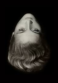 a black and white closeup portrait of a woman's face and hair. the image is taken from a unique perspective, with the subject's head upside down. the woman's face is serene and expressionless, her features softly illuminated against the dark background. her hair is styled in soft waves, typical of the 1930s era. the overall aesthetic is classic, elegant, and subtly mysterious. the monochrome palette enhances the vintage feel and draws attention to the textures of both the skin and hair. the image evokes a sense of timeless beauty and classic hollywood glamour.