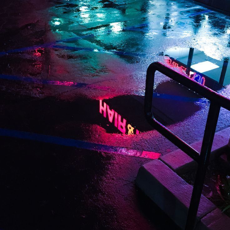 a wet, neonlit urban street at night. the image is composed of a wet, black pavement with a pink and blue neon sign reflecting on the surface. a metal railing runs diagonally across the image, adding depth and texture. the colors are saturated and vibrant, creating a sense of energy and movement. the overall feeling is one of urban vibrancy and mystery.