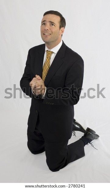 a man in a suit is kneeling on the floor, praying or pleading. he is looking up with a sad and worried expression on his face. he is wearing a black suit with a yellow and black striped tie. his black shoes are visible at the bottom of the frame. the background is white.