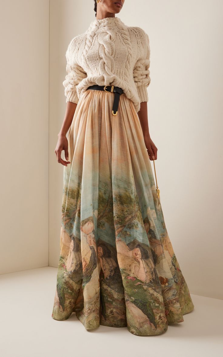 a woman is posing in a white longsleeved sweater and long beige skirt with a floral print.  she is wearing a black belt with a gold buckle. her hair is pulled back off her face. she is standing on a light background.