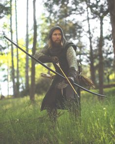 a man dressed as a medieval archer stands in a forest, aiming a bow and arrow. he wears a hooded tunic and leather armor. the image evokes a sense of adventure and fantasy. the lighting is natural and the perspective is at eye level.