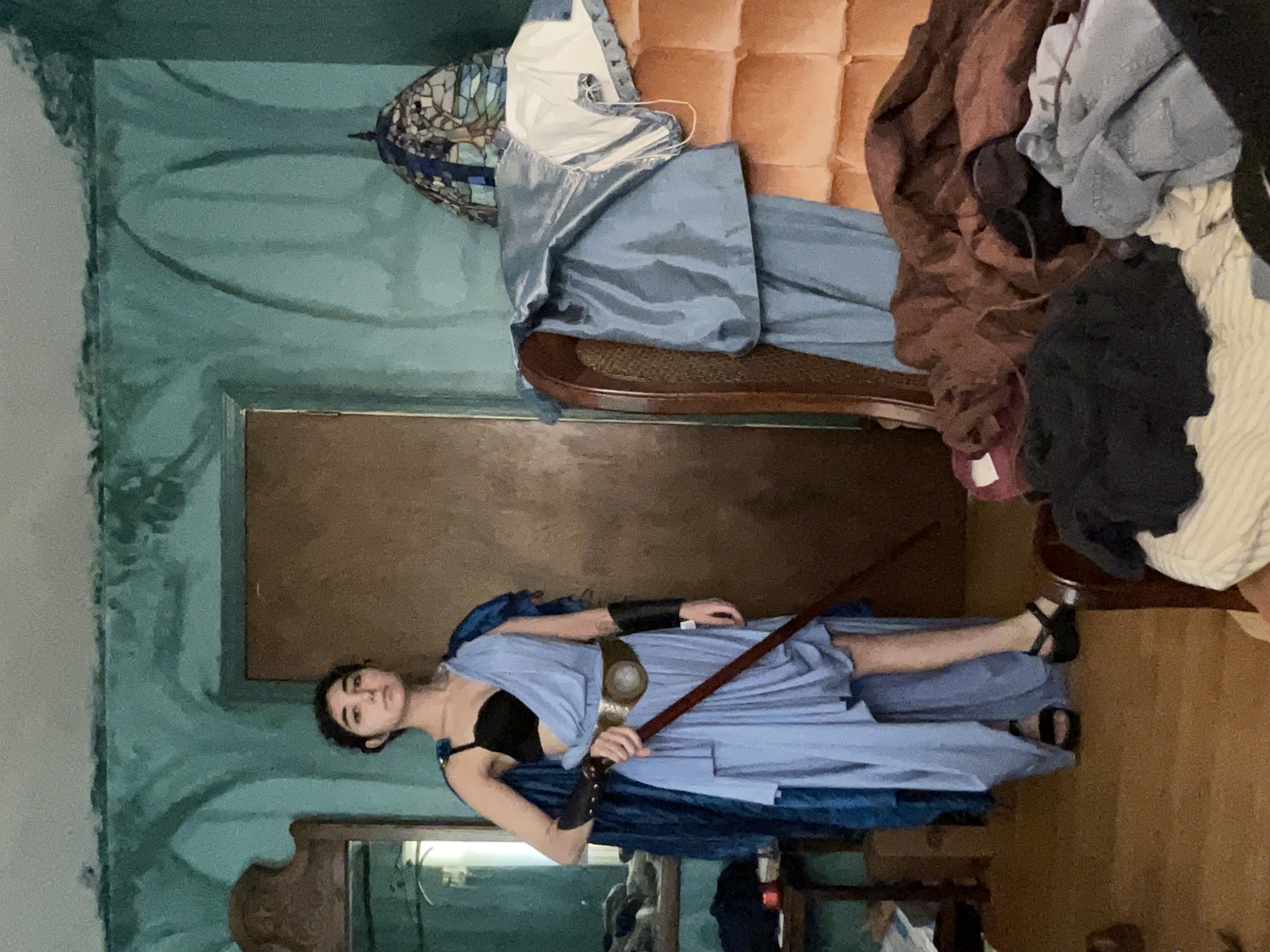 a woman is captured in a full shot, standing in a cluttered bedroom.  she's dressed in a light blue, draped costume reminiscent of ancient greek attire. the dress is asymmetrical, falling to the ground in the back and shorter in the front. she carries a long staff, possibly a prop weapon, and wears dark sandals. her expression is serious and she appears to be posing for a picture.  the room itself is messy, with clothing piled on an armchair and the general environment suggesting a livedin, possibly older house, with teal painted walls. a vintagestyle armchair and a partially visible mirror are visible in the background.  the overall atmosphere blends elements of costuming and everyday life.