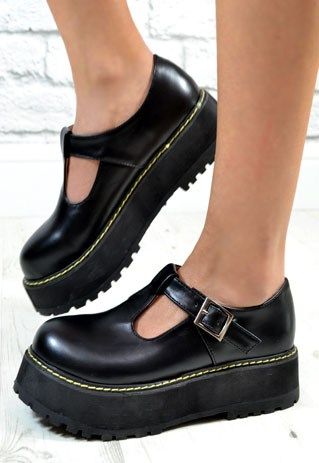 a pair of black platform tstrap shoes are shown on someone's feet, one in front of the other. the shoes have a thick black sole, a shiny black upper with a tstrap design, and a buckle on the strap. the background is a blurry brick wall and a wooden floor.