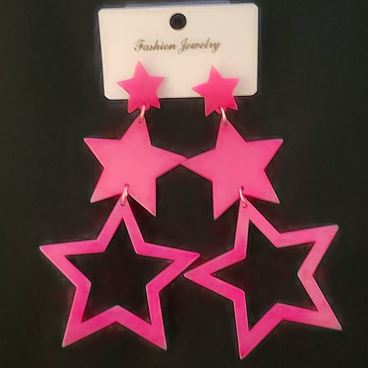 two pairs of pink star earrings hanging from a tag that says "fashion jewelry" are shown on a black background. the earrings are made up of a smaller, solid pink star attached to a larger, hollow star.