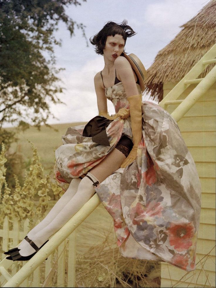 a woman wearing a floral dress, gloves and stockings is sitting on a yellow ladder in an outdoor setting. there is a wooden fence behind her. she is holding a straw hat in her hand. she is looking at the camera with a whimsical expression. the image is a vintage style portrait.