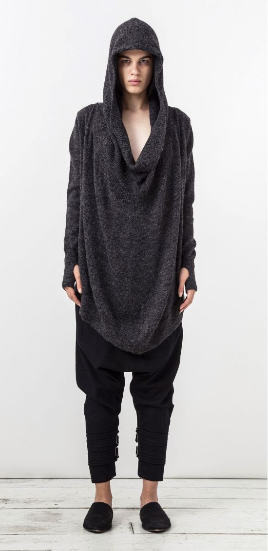 a fulllength studio shot shows a tall, thin man with light skin, wearing a dark gray hooded sweatshirt and loose black pants with rolledup cuffs, standing against a white background. the sweatshirt has a deep cowl neck and long sleeves with thumb holes. the pants are baggy and slightly cropped, with the cuffs rolled up and secured with straps. he is wearing black slippers. the man faces forward with a serious expression. the background is a plain white wall and floor.