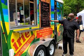 a man is standing next to a bright yellow food truck with the word "irie" written on the side. the truck is decorated with green and yellow colors. the man is wearing a black shirt and black pants and has a black face mask on. he is pointing at the menu on the side of the truck. there are other people in the background, and the truck is parked on a street.