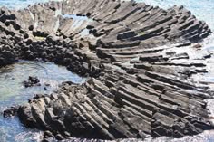 a closeup shot of a rocky shore with black rock formations.  the rocks are stacked in a way that looks like steps or stairs. the water can be seen in the distance, and the rock formations appear to be at the top of a cliff.