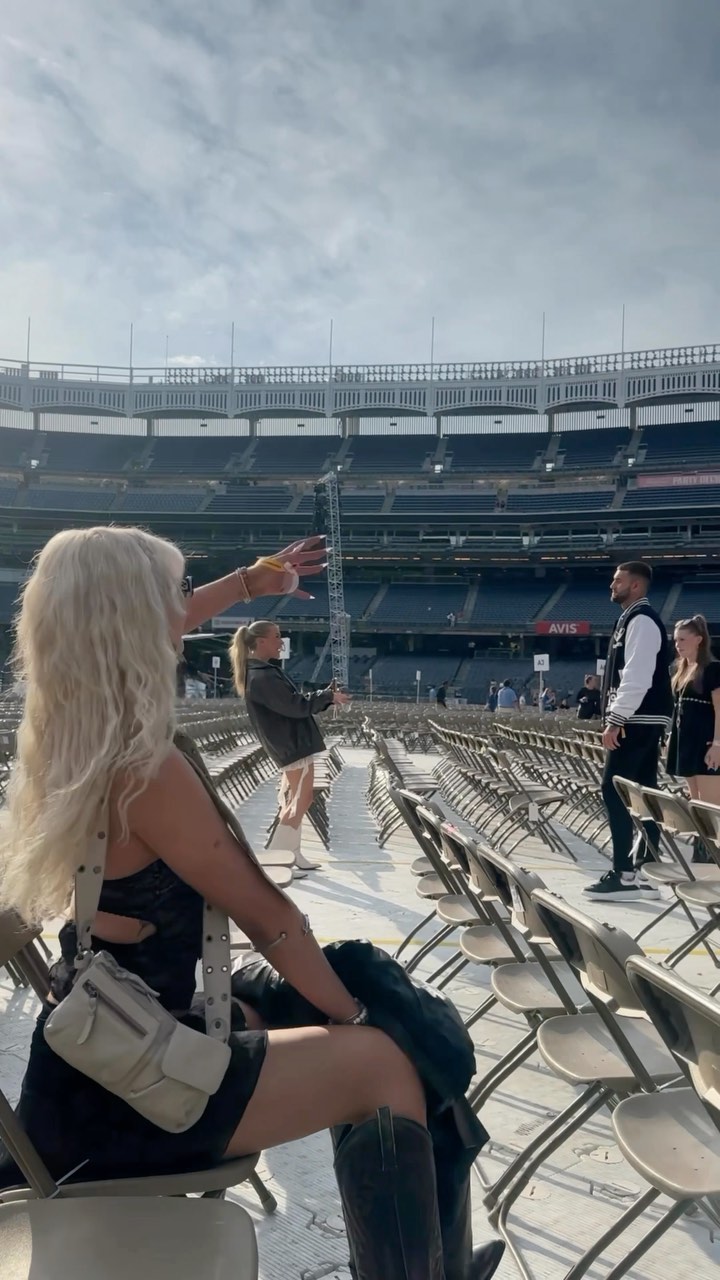 a mediumshot depicts a group of people inside a stadium, likely during or preparing for an event. a blonde woman sits in the foreground, wearing a dark outfit and dark boots, with a lightcolored purse slung over her shoulder. she is pointing her arm out, possibly indicating something to someone. in the middle ground, another woman in a jacket and light pants appears to be adjusting or holding a part of the stage setup. a man in a varsity jacket stands nearby. the stadium is filled with rows of empty seats. the lighting suggests it is daytime.