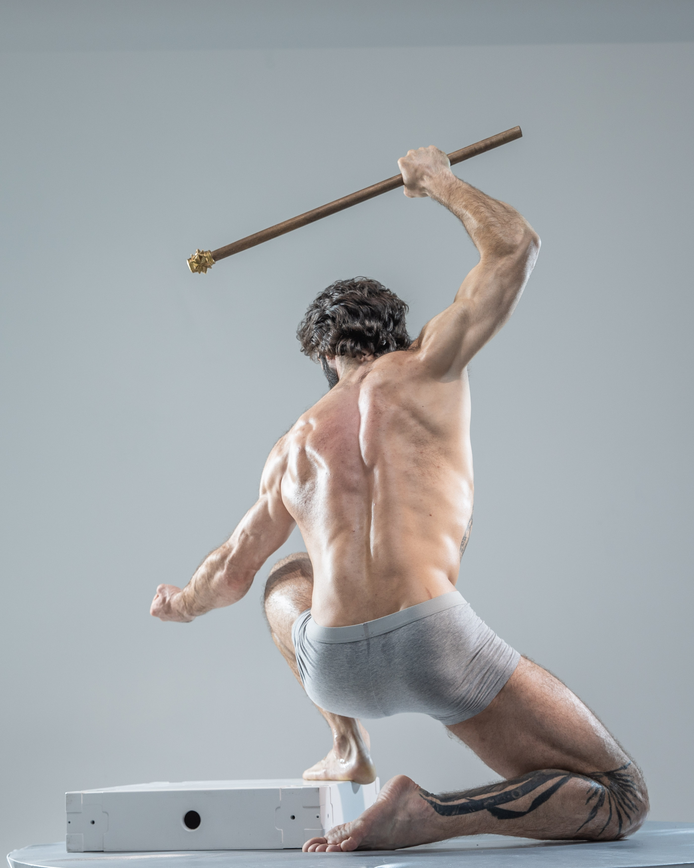 a muscular man with tattoos is posed dynamically against a light gray background. he is holding a staff in his right hand and is wearing gray underwear. his back is to the camera, and his body is angled to the left. he is looking over his left shoulder. the lighting is bright and even, highlighting the man's muscular physique. there is a white cube in front of him.
