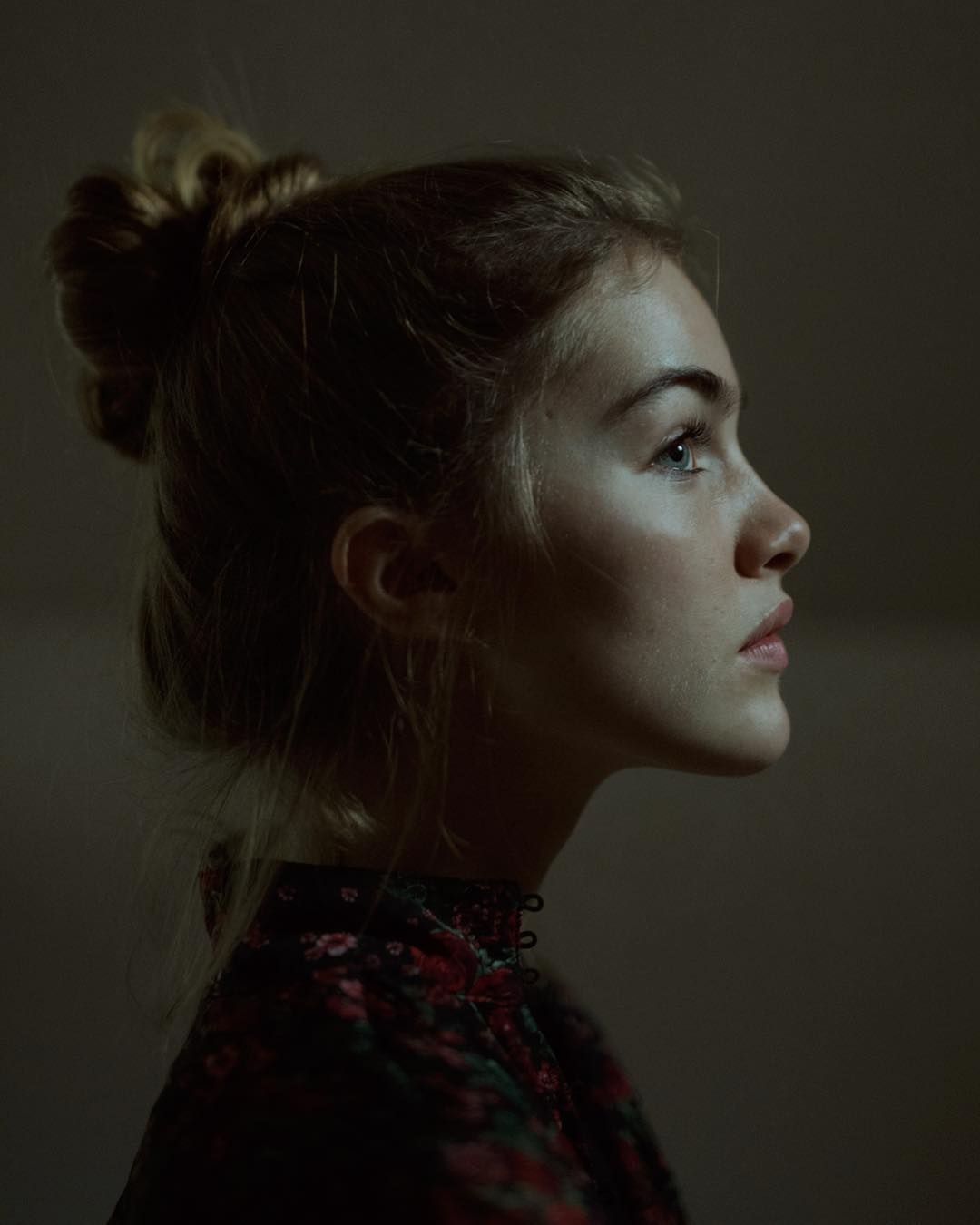 a profile portrait of a young woman with fair skin and light brown hair styled in a bun. she's wearing a dark floral top. her gaze is directed upwards with a thoughtful or contemplative expression. the lighting is soft, highlighting her features against a dark background.
