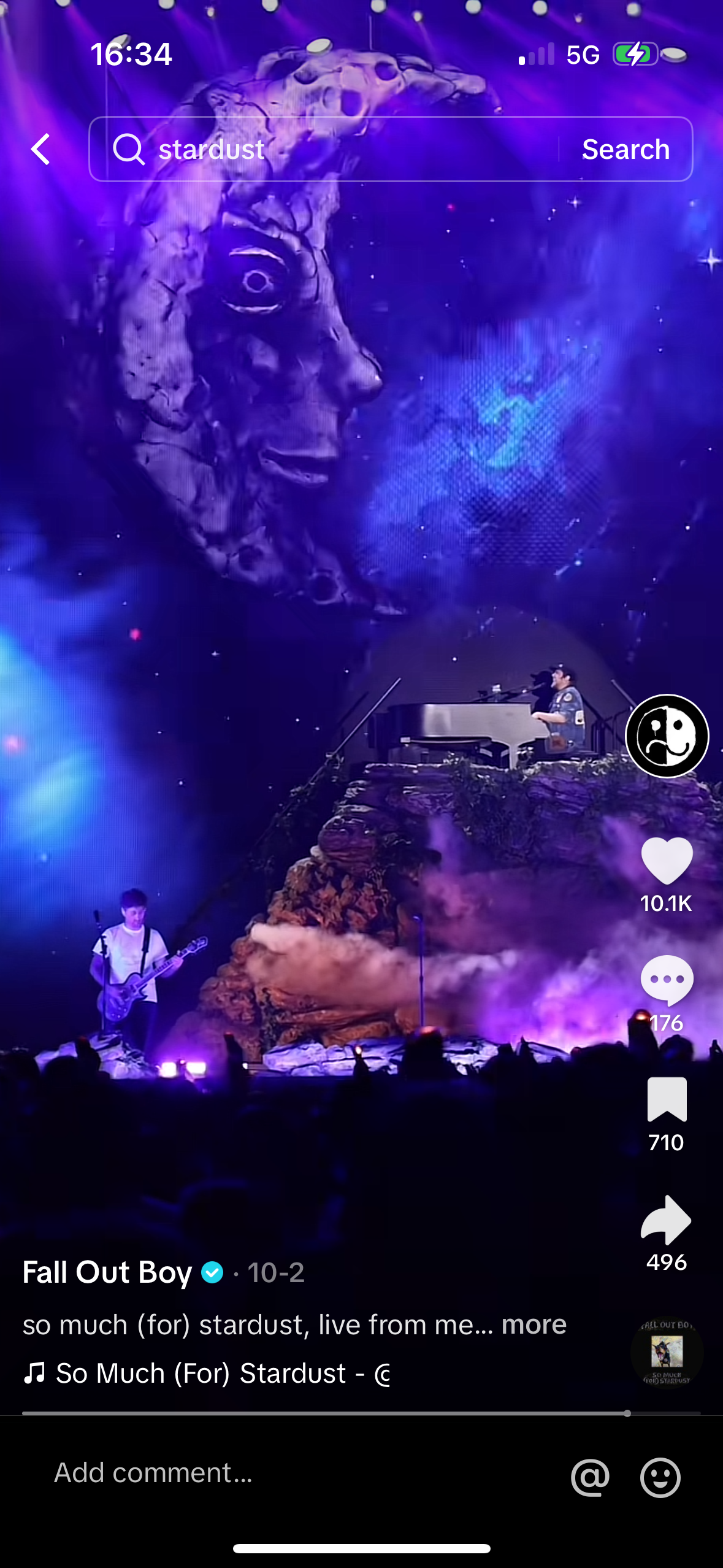 a photo of a live performance of fall out boy, featuring a guitarist with a microphone stand on a stage in front of a crowd of excited fans. the band is performing their song "so much (for) stardust" from their album "so much (for)  stardust" and the photo has been captured from the audience with a cell phone. the photo is captioned with the band name, the song title, and a timestamp for the live performance.  the photo is also filled with some emoji's like a heart, a thinking emoji, a thumbs up emoji and a sharing emoji.