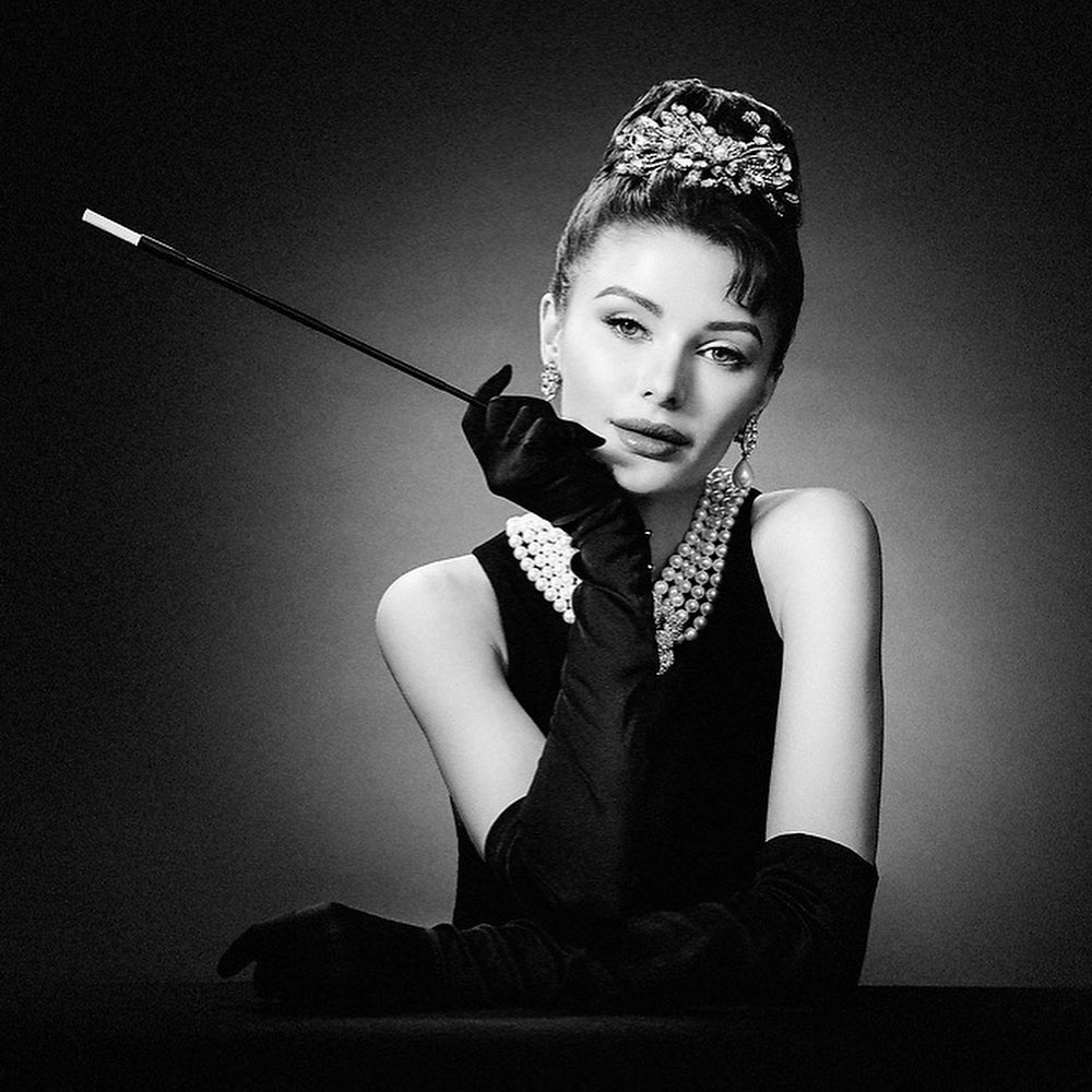 a black and white portrait of a woman with elegant style. she is wearing a black dress, long black gloves, a pearl necklace, and a jeweled hair accessory. she is holding a cigarette and staring at the camera with a slightly knowing smile. the photo has a retro feel and captures the glamour and sophistication of a bygone era.