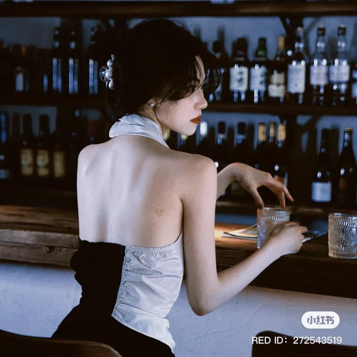 a woman sits at a bar with her back to the viewer. she is wearing a white and black halter top dress. her hair is up and she has a pearl hair clip. there are bottles behind her on the shelves and two glasses on the counter.