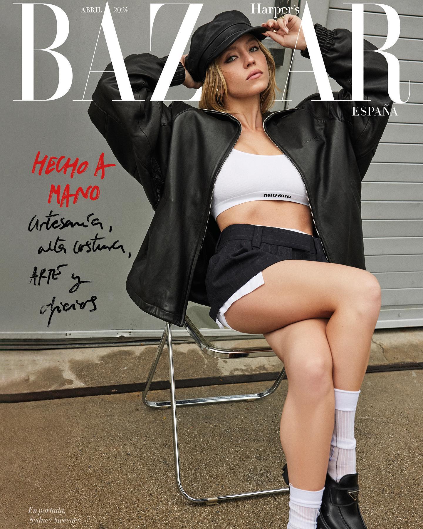 the image is a cover of harper's bazaar spain's april 2024 issue, featuring actress sydney sweeney.  sweeney is seated on a metallic folding chair outdoors.  she's wearing a black leather jacket, a white miu miu sports bra, black shorts, white socks, and prada shoes. the overall aesthetic is a blend of street style and high fashion, consistent with the magazine's focus on "handmade artistry and haute couture." the background is a simple grey wall, with spanish text emphasizing the handcrafted nature of the featured fashion.