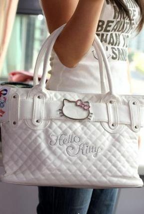 a person is holding a large, white, quilted hello kitty purse. the purse has a white strap across the middle with metal accents and a hello kitty logo with a pink bow. the purse is embroidered with the words "hello kitty." the person is wearing a white top and blue jeans.