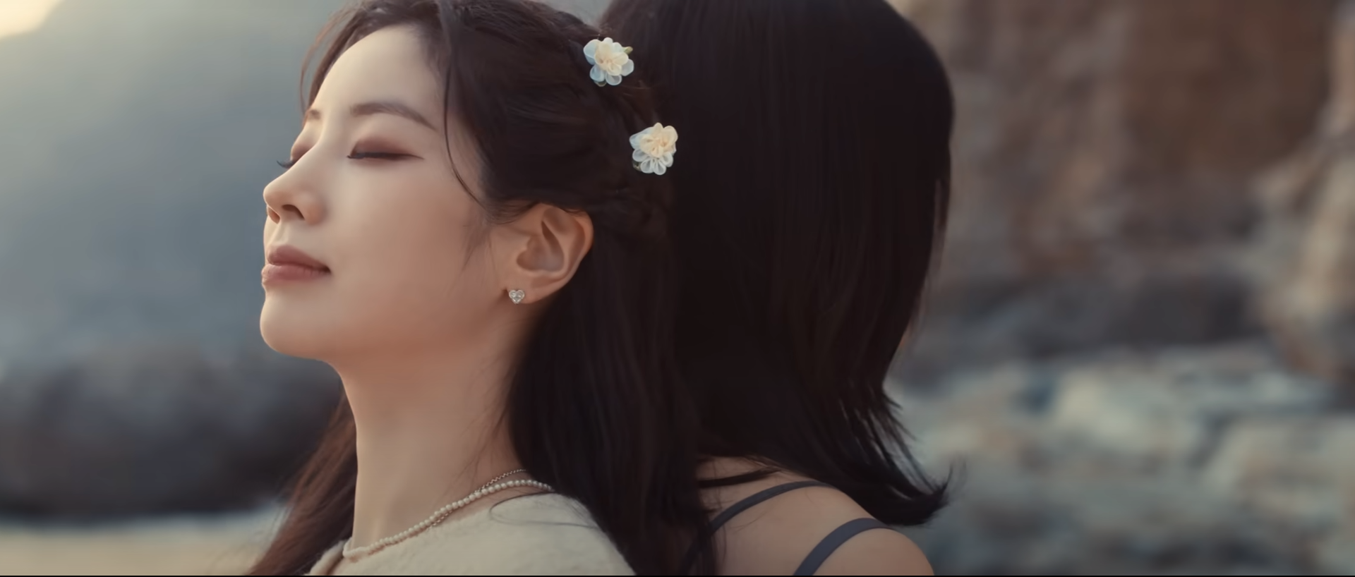 the image showcases two women embracing on a rocky beach at sunset. the focus is on the woman in the foreground, with her eyes closed and a serene expression. her hair is adorned with small, delicate flowers. the background features a blurry seascape and rocky cliffs, bathed in the warm glow of the setting sun. the overall mood of the image is one of peace, intimacy, and connection.