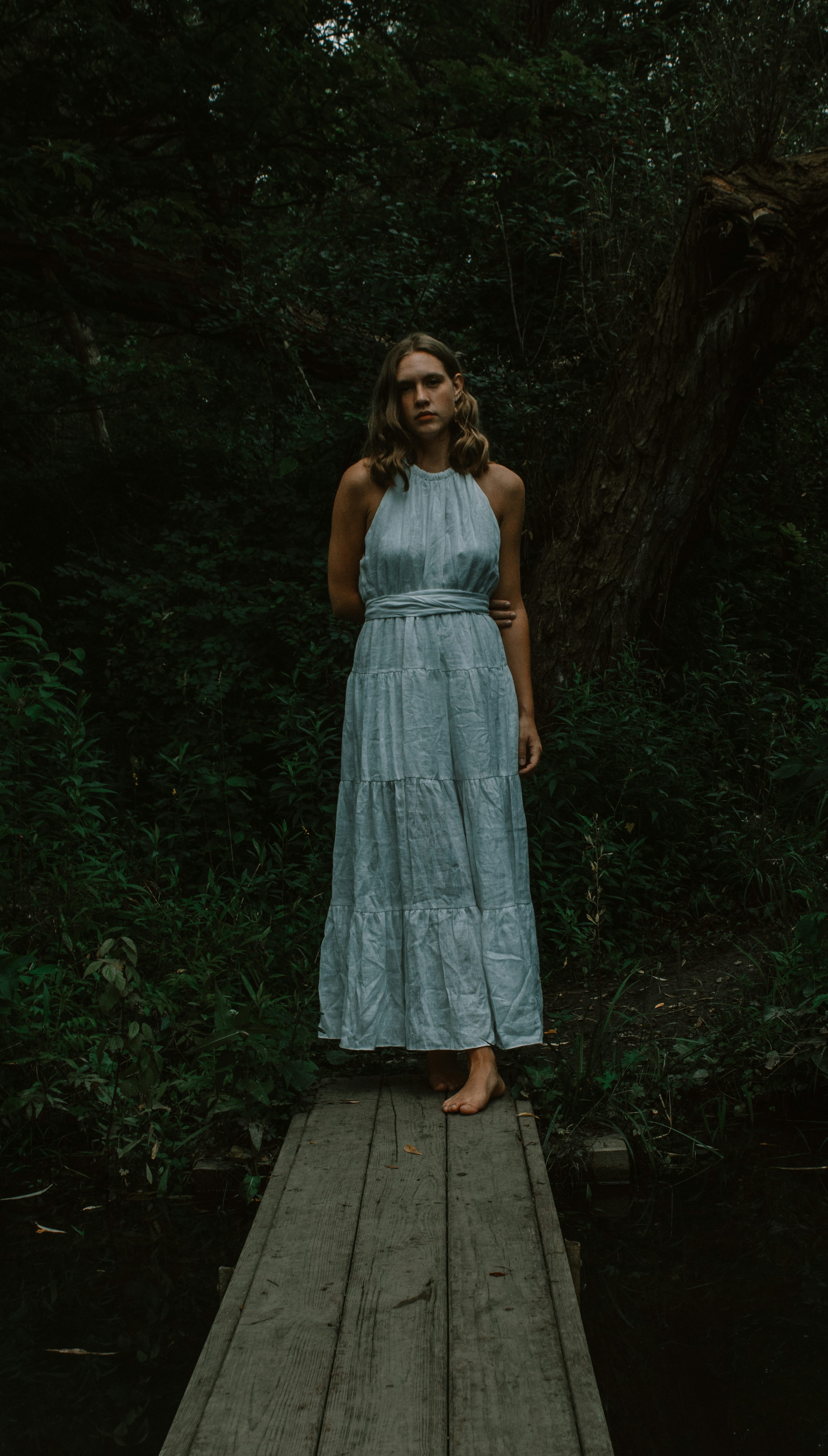 a young woman in a light blue tiered maxi dress stands barefoot on a wooden walkway in a dark forest. the atmosphere is moody and atmospheric, with shadows and greenery surrounding her. she appears serene and calm, lost in thought. the image evokes a feeling of mystery and solitude.