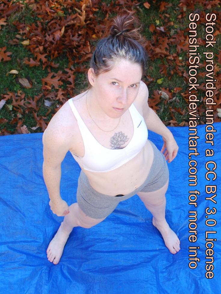 a woman in a sports bra and athletic shorts stands on a blue tarp in a fighting stance. she is looking up at the camera, and there are fallen leaves on the ground around her. the image is taken from a high angle, giving the viewer a bird's eye view.