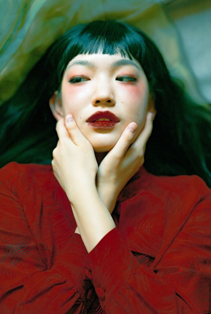 a portrait of a young woman with dark hair and red lips. she is wearing a red dress and her hands are resting on her cheeks. the background is green and the photo has a vintage feel to it.