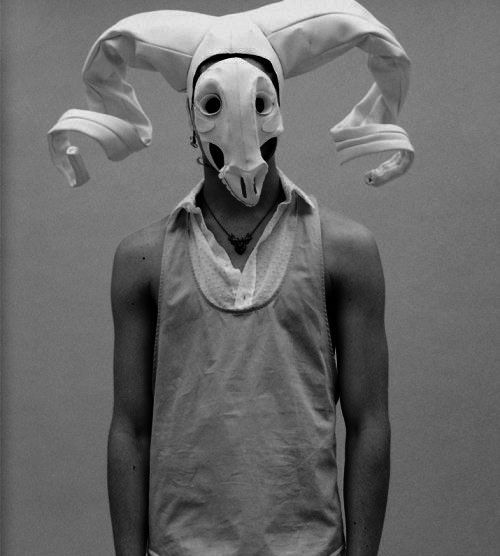 a black and white portrait of a man wearing a white, surreal mask with horns. the mask is made of fabric and covers the man's face, with the only part visible being the eyes, which are obscured by the mask's design. the man is wearing a white tank top, and his hair is hidden beneath the mask. the background is a plain white wall. the overall effect of the image is dramatic and artistic, with a strong sense of mystery. the man's expression is hidden by the mask, adding to the enigmatic quality of the image. the bold and expressive nature of the mask and the man's outfit suggests a conceptual or avantgarde aesthetic.