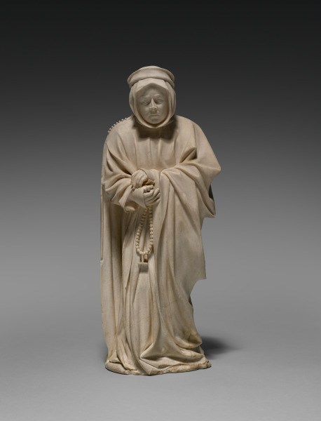the image is a photograph of a marble sculpture depicting a woman wearing a robe and a headdress. she is holding a rosary in her hands. the sculpture is set against a plain grey background.