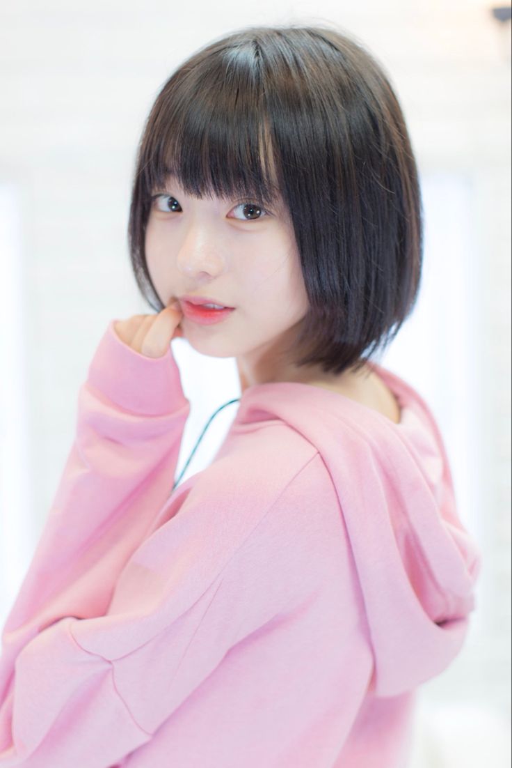 an asian girl with black hair is wearing a pink hoodie while looking at the camera and smiling. she is posing in front of a white background.