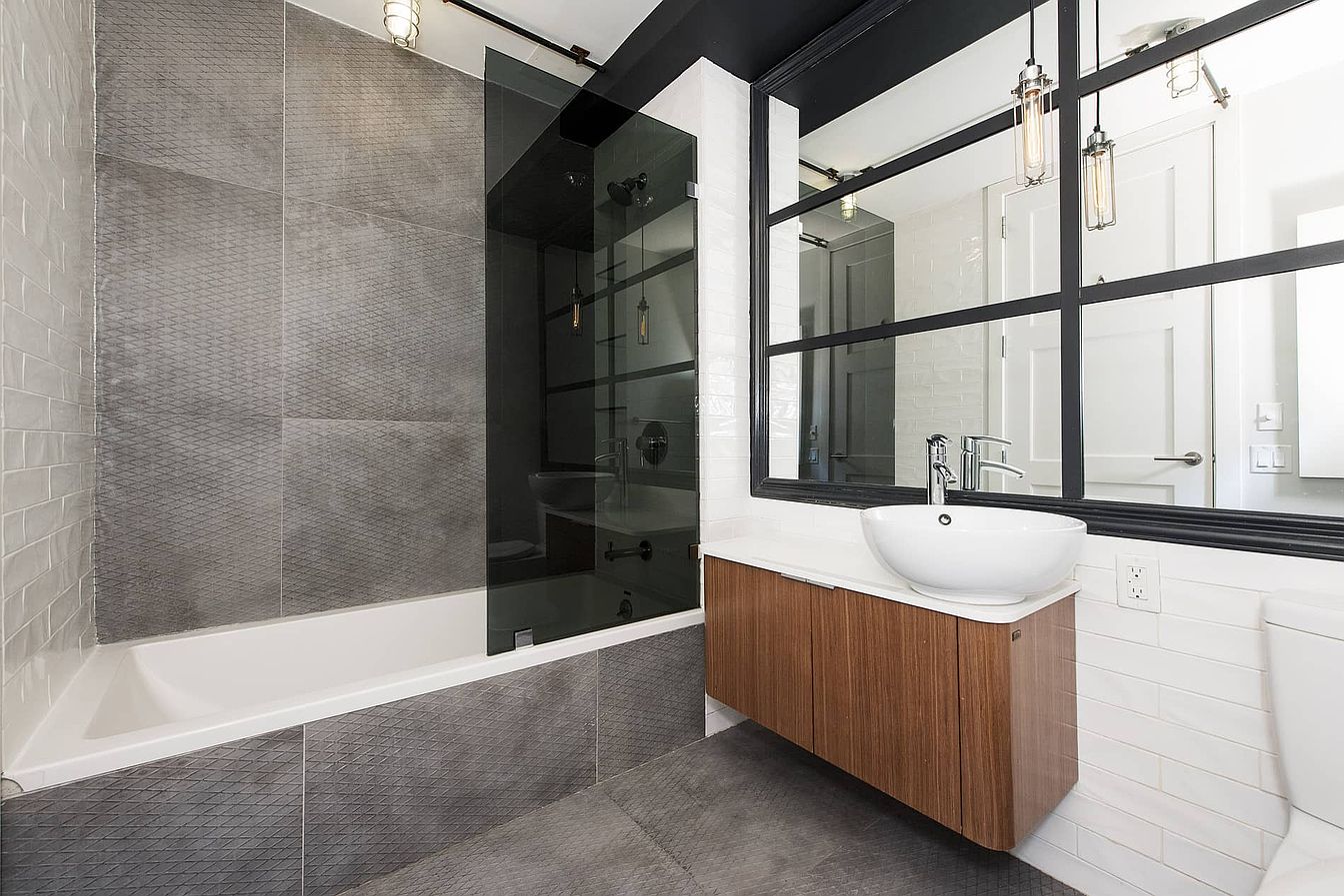 a modern bathroom with gray tile walls and a white tub. the shower is behind a glass door, and the vanity has a wooden cabinet and a white sink. the mirror above the vanity has a black frame and is surrounded by white subway tiles. the lights are hanging from the ceiling and are reflected in the mirror.