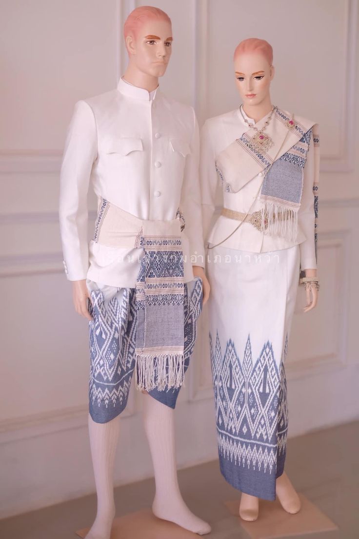 a thai couple in traditional wedding attire. the groom is wearing a white longsleeved shirt with a high collar and a blue and white patterned sarong. the bride is wearing a white dress with a blue and white patterned sash and a white shawl. they are both standing in front of a white wall. the image is elegant and portrays the beauty of traditional thai wedding attire.