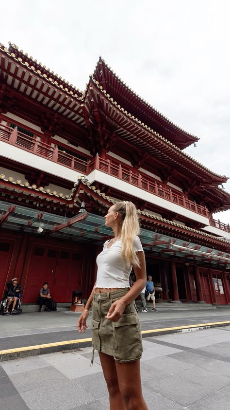 a young woman in a white shirt and olive green mini skirt stands in front of the buddha tooth relic temple in singapore. the temple's intricate red and white architecture dominates the background, and the woman is posed looking up towards the building.  the scene is shot from a low angle, emphasizing the height and grandeur of the temple. other people are visible in the background but are out of focus, the main subject being the woman and the temple.  the overall mood is one of travel and sightseeing in an urban asian setting.