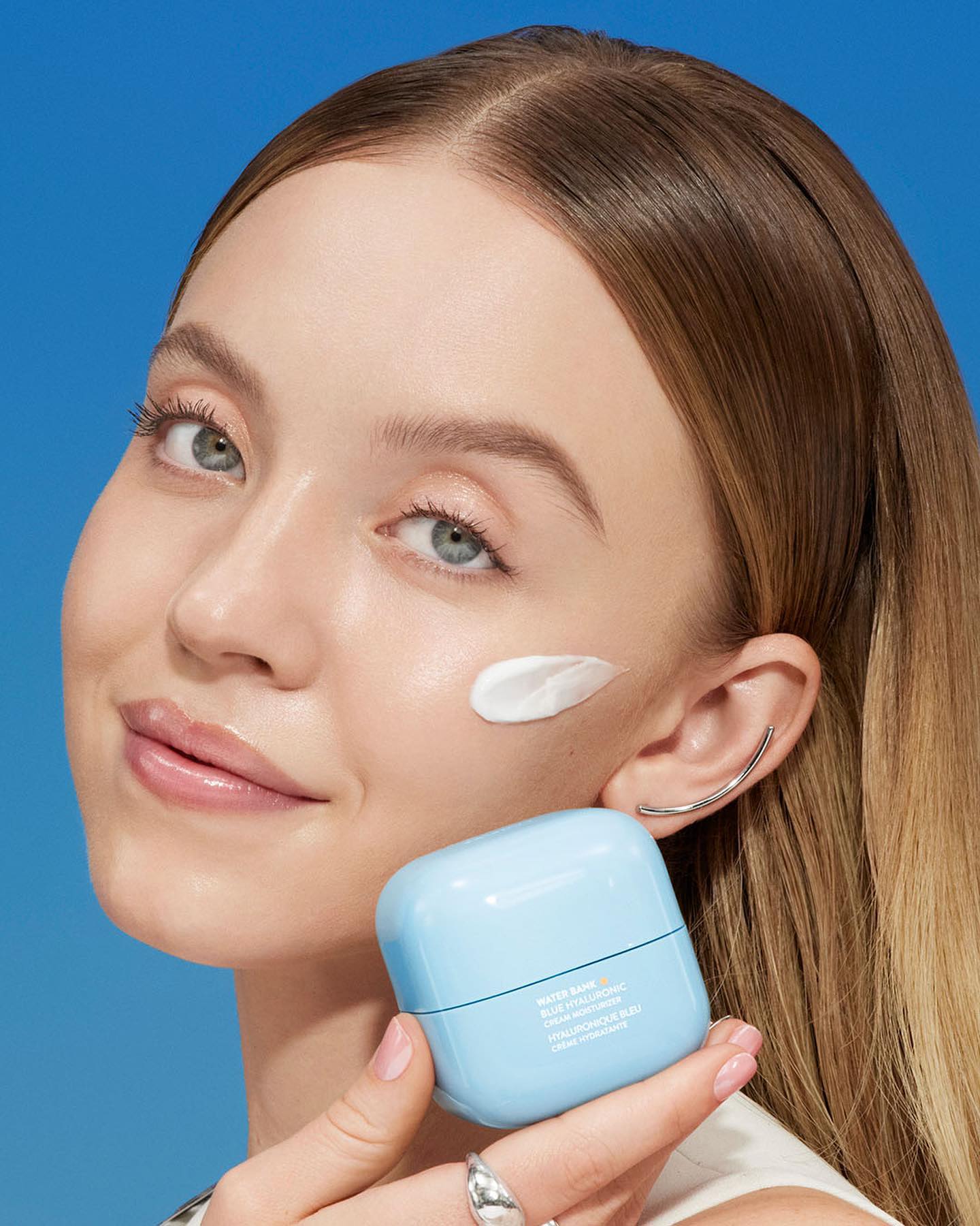 a closeup shot of a young woman with light skin, applying a light blue moisturizer to her cheek.  the moisturizer is in a small, square, light blue jar and the brand name, water bank, is visible on the packaging.  the woman is smiling slightly and looks directly at the camera. the background is a solid light blue color. the overall feeling is clean, fresh, and bright, promoting the skincare product.