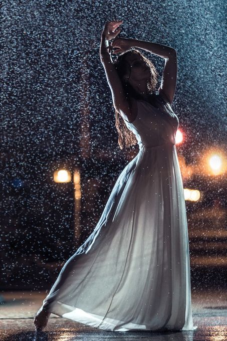 a woman in a white dress is dancing in the rain at night. the city lights are blurred in the background. she is silhouetted against the rain, and her dress is flowing gracefully. the image is romantic and mysterious.