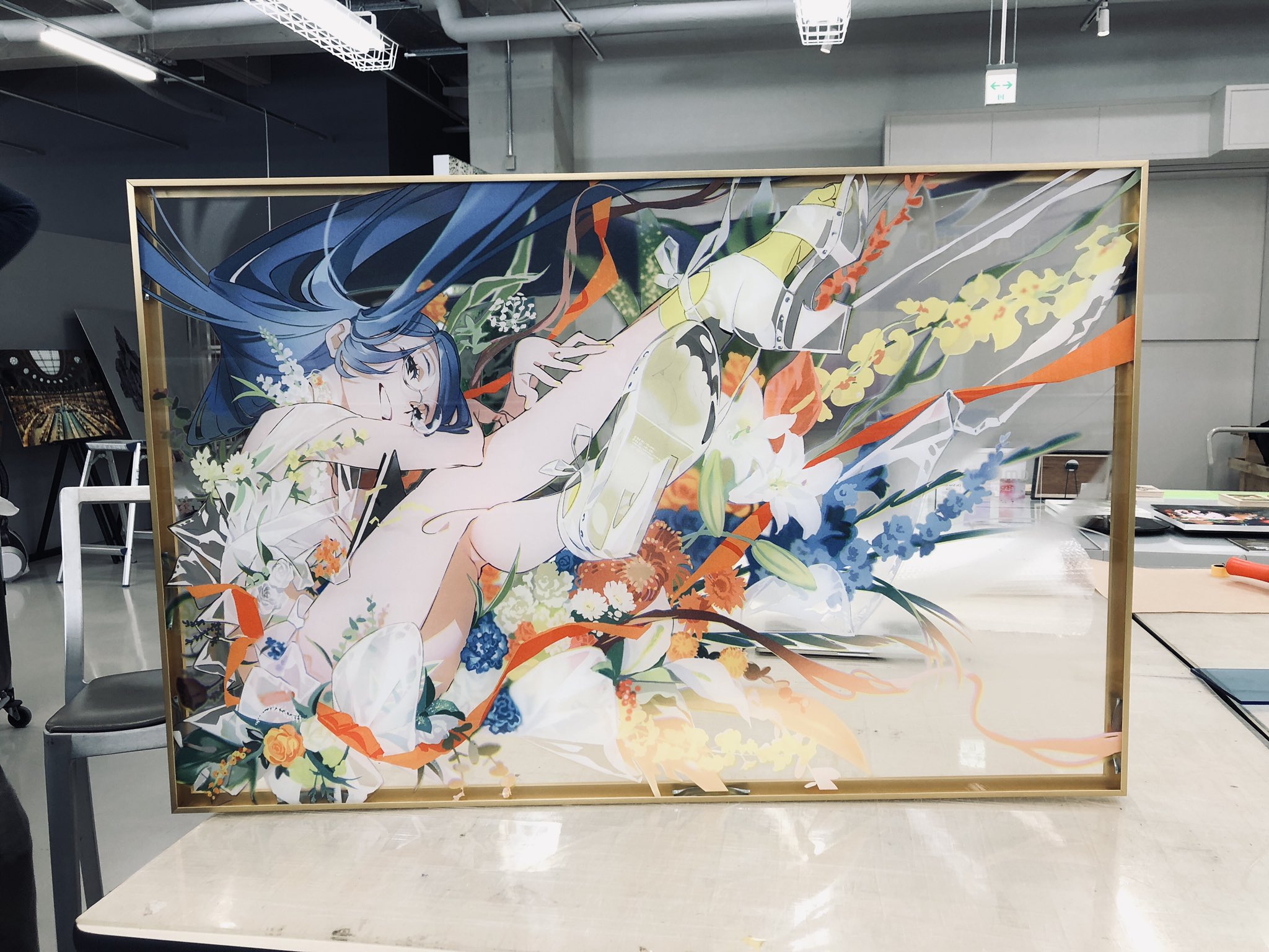 the image showcases a framed acrylic artwork featuring an animestyle female character with blue hair. she's depicted lying amidst a vibrant array of flowers. the artwork is placed on a table within what appears to be an art studio. the setting suggests the piece might be newly finished or in the process of being mounted or displayed.