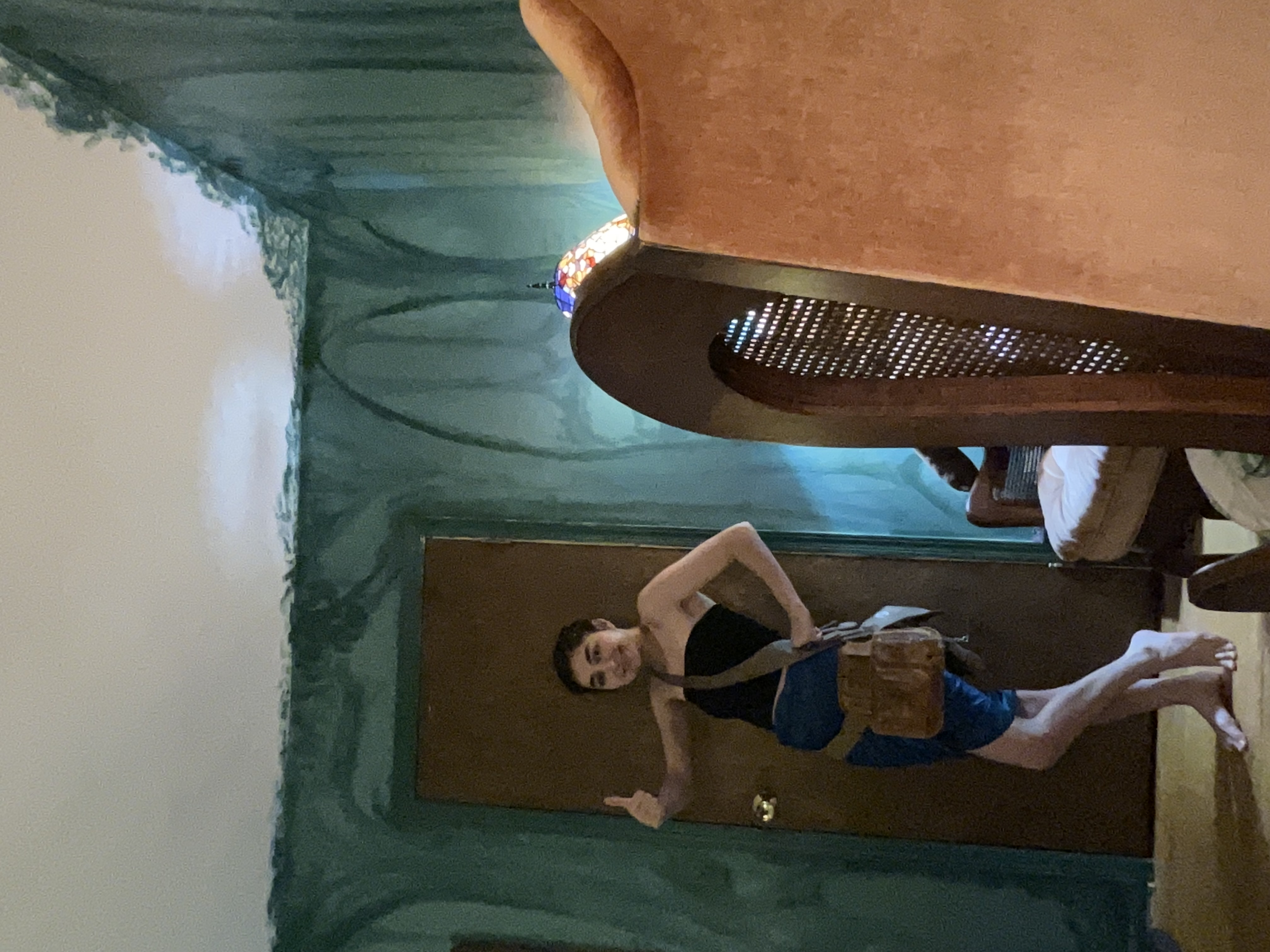 a young woman with dark hair and a light complexion stands in a room with uniquely painted walls, giving a thumbsup. she's wearing a short darkcolored skirt and a brown messengerstyle bag. the walls are painted in shades of teal and green, featuring a design that resembles stylized trees or vines. a partially visible orange chair with a wooden frame is in the background to the right.  the overall lighting in the room suggests it may be an evening shot.