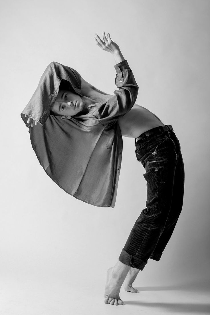 a black and white fullbody shot of a male contemporary dancer in midmovement. he is wearing a loose, oversized shirt and jeans, his torso bare. his body is arched in a dynamic pose, arms extended, one hand raised high, the other gently resting on his hip. his expression is focused, his feet barefoot. the background is a plain, lightgrey studio setting. the overall impression is one of artistic movement, grace, and elegance.