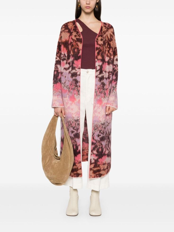 a woman in a colorful floral print cardigan, white pants, and white boots poses with a brown leather bag.