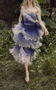 a young woman in a beautiful blue and white tulle dress stands in a forest, looking thoughtfully into the distance. the dress is flowy and ethereal, with delicate ruffles and layers. the sunlight filters through the trees, creating a soft and magical atmosphere.