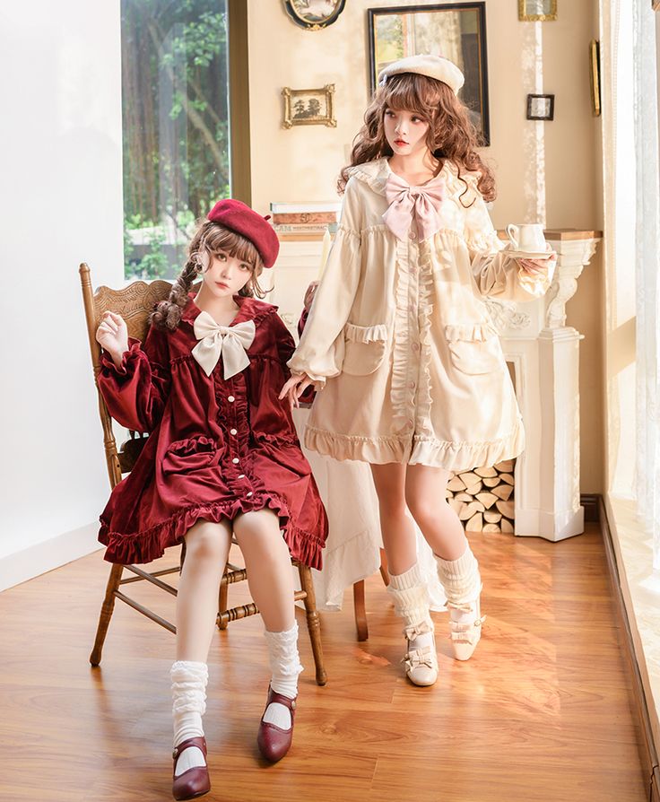 two women are posing in a room. they are both wearing lolita fashion dresses and berets. the woman on the left is wearing a red dress and the woman on the right is wearing a cream dress. they are both smiling and looking at the camera.