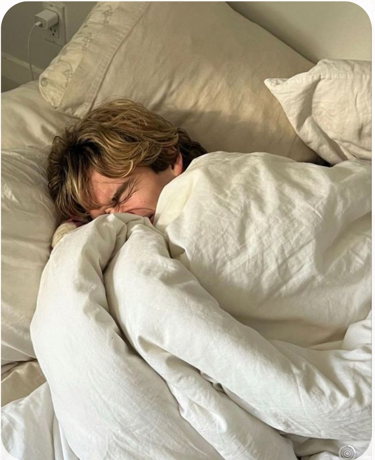 a young man with tousled blonde hair is sleeping soundly in a bed. he is snuggled deeply under white bedsheets, with his face partially obscured. the sunlight streams softly into the room, creating a warm and inviting atmosphere. the overall feeling of the image is one of comfort and tranquility.
