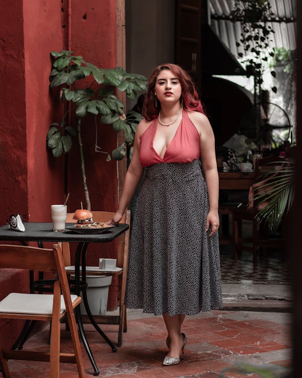 a woman with long red hair is standing outdoors at a cafe. she is wearing a coral halter top and a black and white polka dot midi skirt. she is also wearing heels. she is looking directly at the camera with a confident expression. the setting is a patio with red walls and lush plants. there is a table in front of her with food on it.