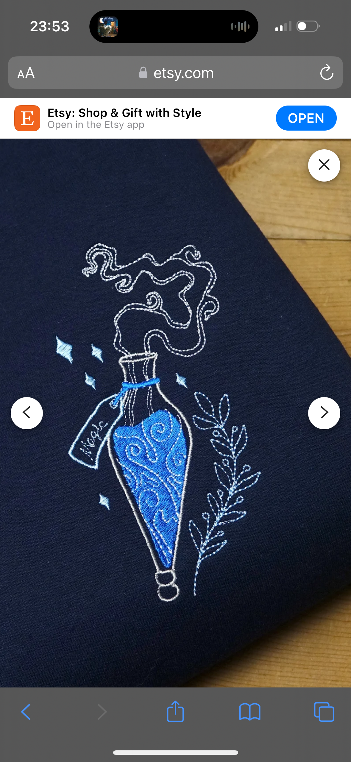 a closeup image of a dark blue fabric with a white embroidered potion bottle with swirling blue liquid and white smoke coming out of the top. the bottle has a tag that reads "magic" on it, and there are white flowers and other designs around it. the fabric is folded, and the wooden table is visible in the background.