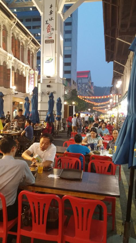 the image shows a bustling night market or hawker centre in singapore.  the scene is vibrant and lively, with numerous people sitting at red plastic chairs and tables, enjoying meals.  the setting is outdoors, amongst traditionallooking buildings, some with colonial architecture, and modern highrises visible in the background.  umbrellas provide shade, and there are lanterns and lights hanging overhead, adding to the atmosphere. the scene is very crowded, indicating a popular and busy food area.  chinese characters are visible on some signs.  the overall impression is one of a lively, cultural, and authentic food experience.