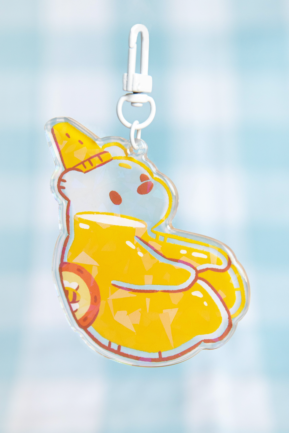 closeup shot of a cute acrylic keychain featuring a cartoon polar bear wearing a yellow ice cream cone hat and sitting in a yellow inflatable ring.  the keychain is transparent, showing the image printed on the acrylic. the keychain has a white plastic clip at the top. the background is a blurry light blue.