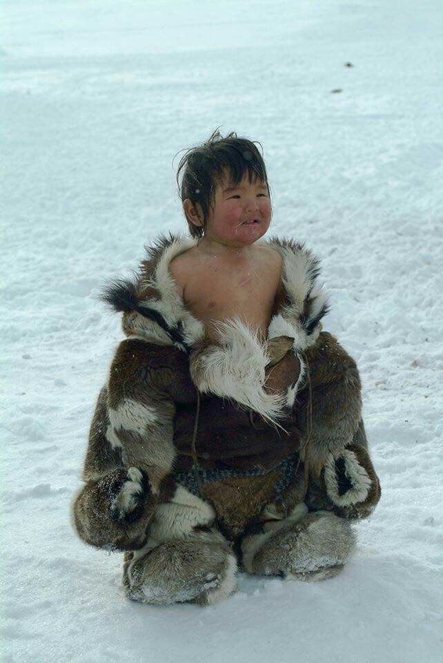 a young inuit child sits in the snow wearing a traditional fur parka. the child has a happy expression and is partially undressed, suggesting resilience to the cold. the background is a vast expanse of white snow, emphasizing the arctic environment.
