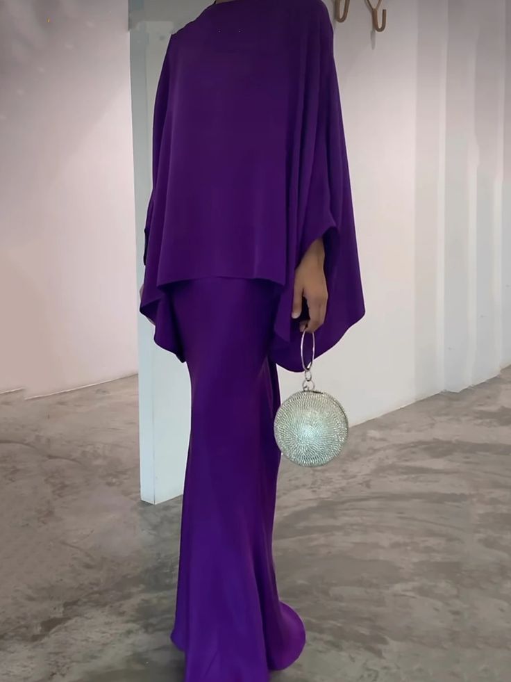 the image shows a woman wearing a floorlength, deep purple dress. the dress has wide, flowing sleeves and a modest, somewhat aline silhouette.  it appears to be made from a smooth, possibly silky or satinlike fabric.  the dress is paired with a silver, sparkly, round clutch bag held in the model's hand. the setting is a minimalist, lightcolored room with a plain background.  the overall style is elegant and contemporary.