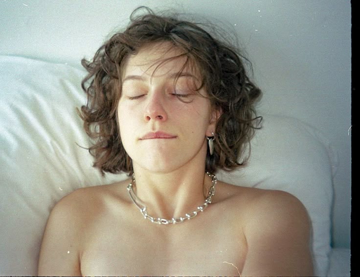 a woman with curly hair is lying on a pillow with her eyes closed. she is wearing a silver necklace and earring. her shoulders are bare.