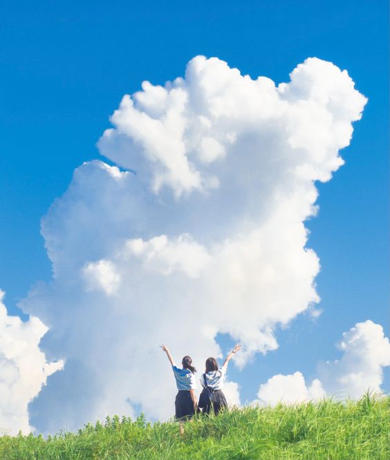 two girls in school uniform are standing on a grassy hill with their arms raised in the air. they are looking up at a large, white cumulus cloud in the blue sky. the sun is shining brightly, and the scene is full of joy and happiness.