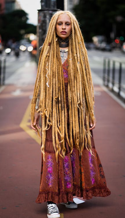 a full shot of a young woman with long blonde dreadlocks stands confidently on a city street. she's wearing a long, brown bohemian maxi dress with purple and pink floral embroidery.  her style is bohemian or hippie, accessorized with a necklace. she wears white sneakers and her expression is serious and direct. the background is blurred, showing a city street with cars and buildings. the overall lighting suggests it is daytime.