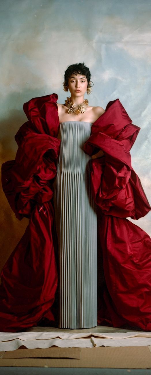 a woman stands in a studio wearing a gray pleated dress with large red puff sleeves and matching decorations. she has short dark hair and is wearing gold earrings and a matching necklace. the backdrop is a painted sky. the woman is the focus of the shot.