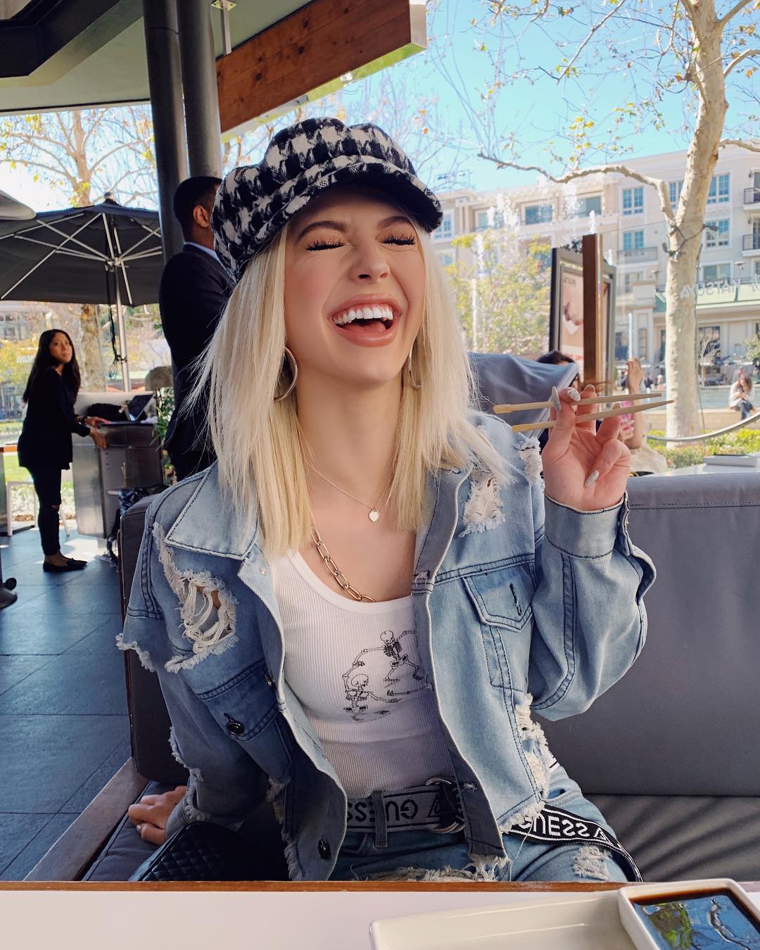 a woman with blonde hair is seated outdoors at a table, laughing and holding chopsticks in her hand. she is wearing a patterned hat, a ripped denim jacket over a white tank top, and a "guess" belt. her outfit is completed with jewelry, including hoop earrings and layered necklaces. behind her, the background shows trees, buildings, and other people in a daytime setting.